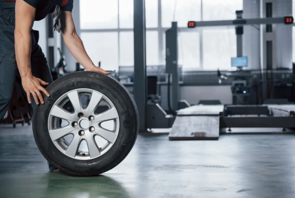 A mechanic in dark coveralls rolls a tire mounted on a silver alloy wheel across the floor of an auto repair shop.