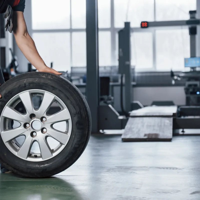 A mechanic in dark coveralls rolls a tire mounted on a silver alloy wheel across the floor of an auto repair shop.