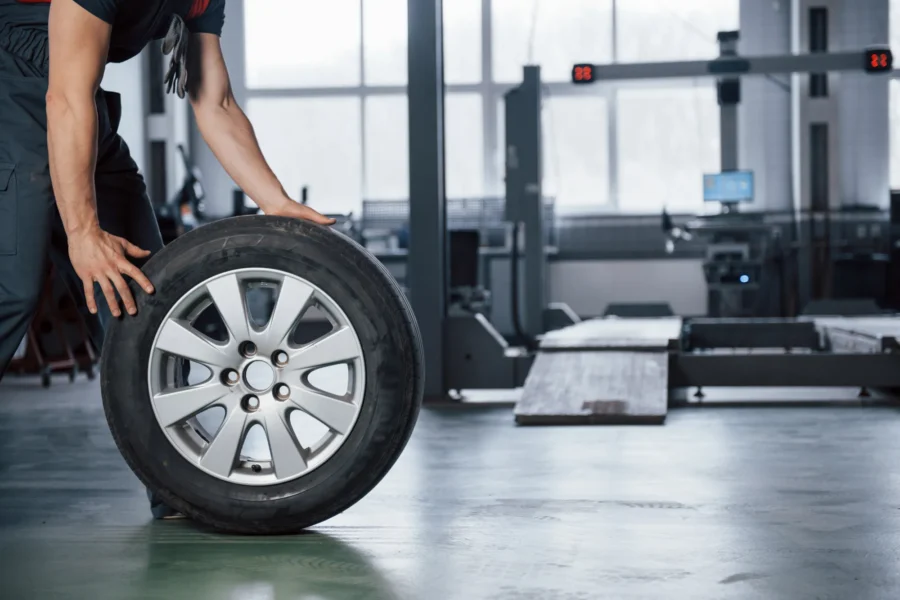 A mechanic in dark coveralls rolls a tire mounted on a silver alloy wheel across the floor of an auto repair shop.
