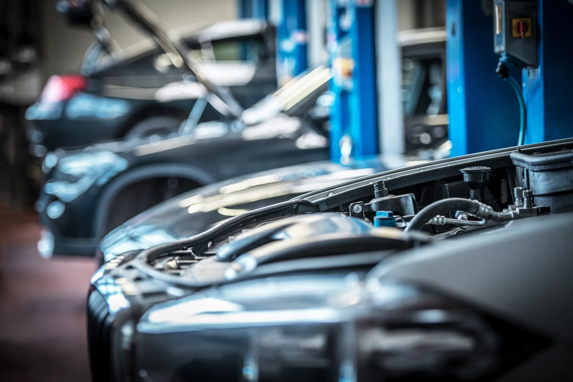 Two cars with their hoods open inside an auto repair shop, with blue vehicle lifts in the blurred background.