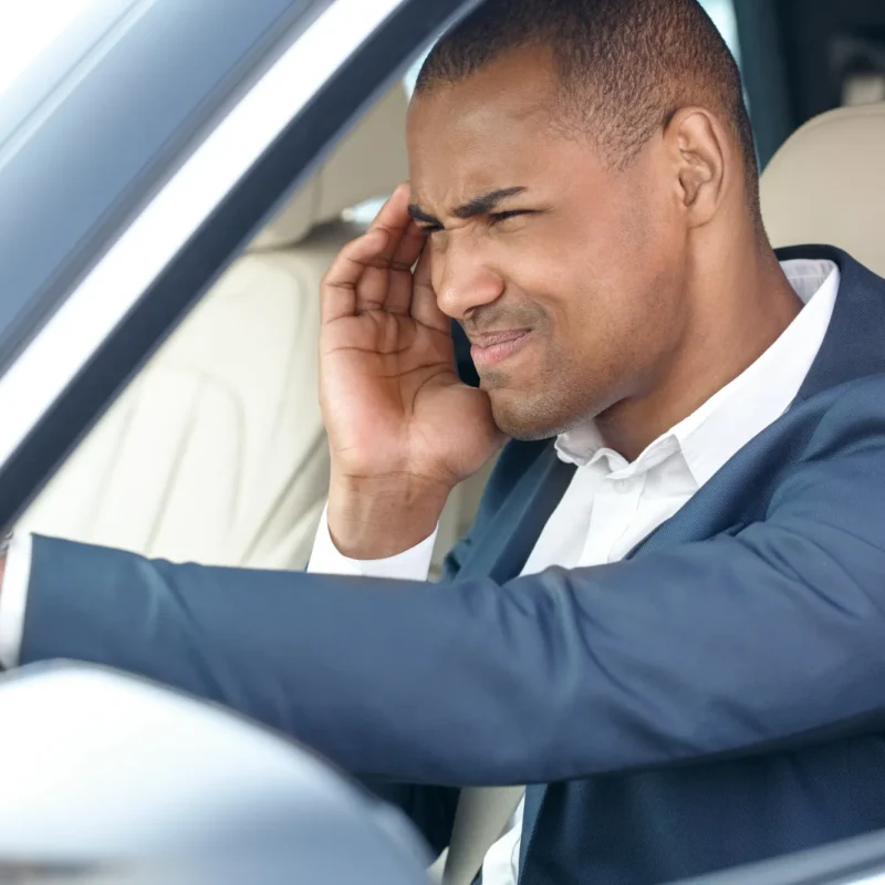 A man in a navy suit sits in a car's driver's seat holding his head in frustration.