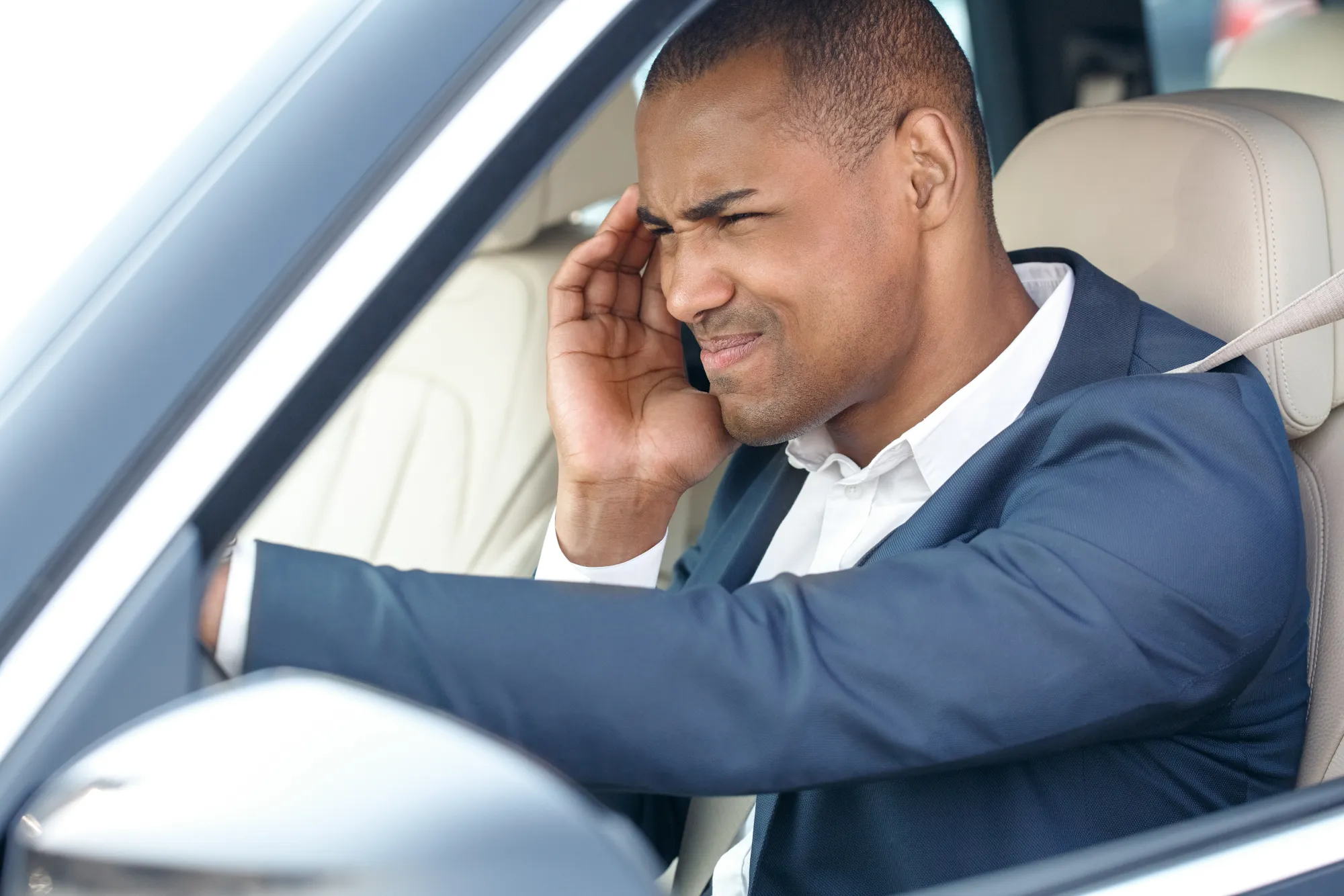 A man in a navy suit sits in a car's driver's seat holding his head in frustration.