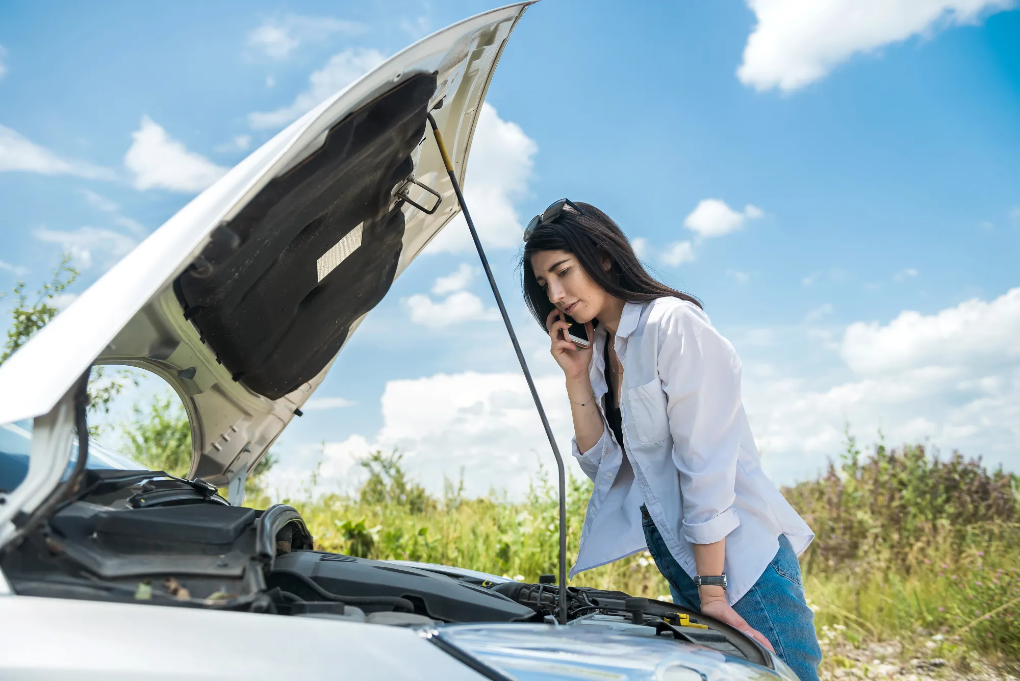 A woman talks on her cell phone while standing next to her white car with its hood raised on a grassy roadside.