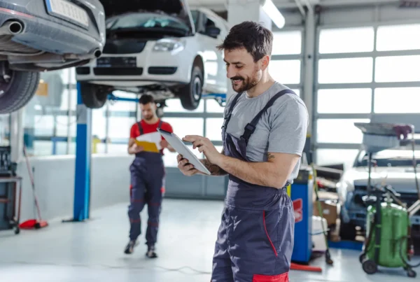 A mechanic in grey overalls looks at a tablet while a coworker with a clipboard stands near a car raised on a lift inside an auto repair shop.