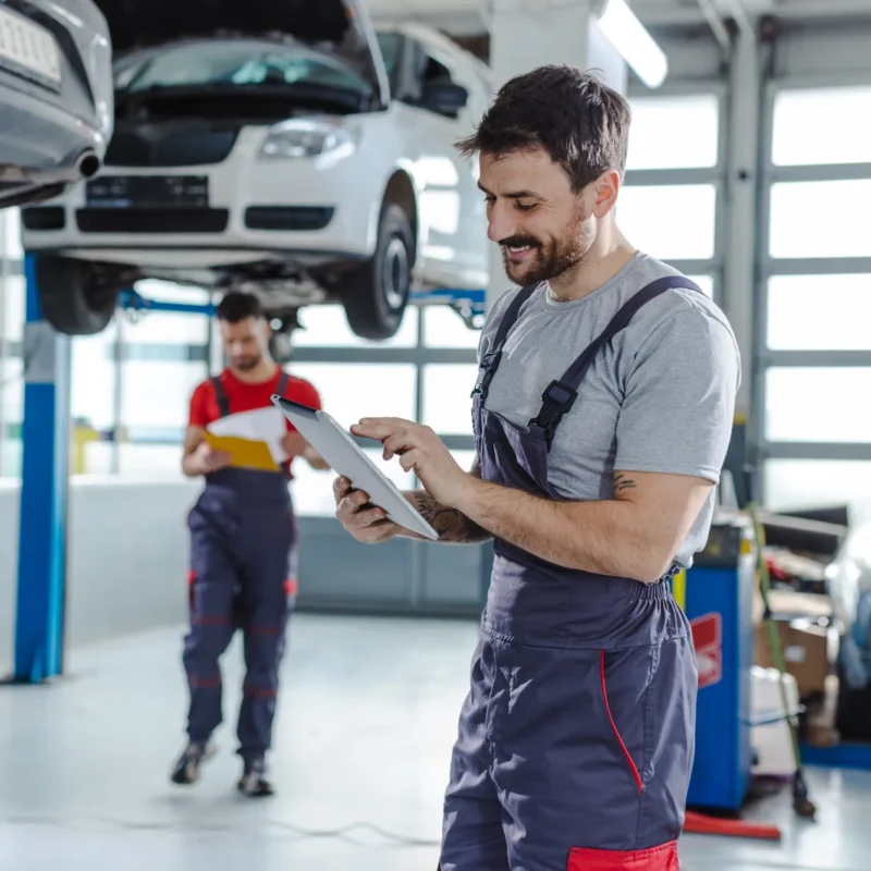 A mechanic in grey overalls looks at a tablet while a coworker with a clipboard stands near a car raised on a lift inside an auto repair shop.