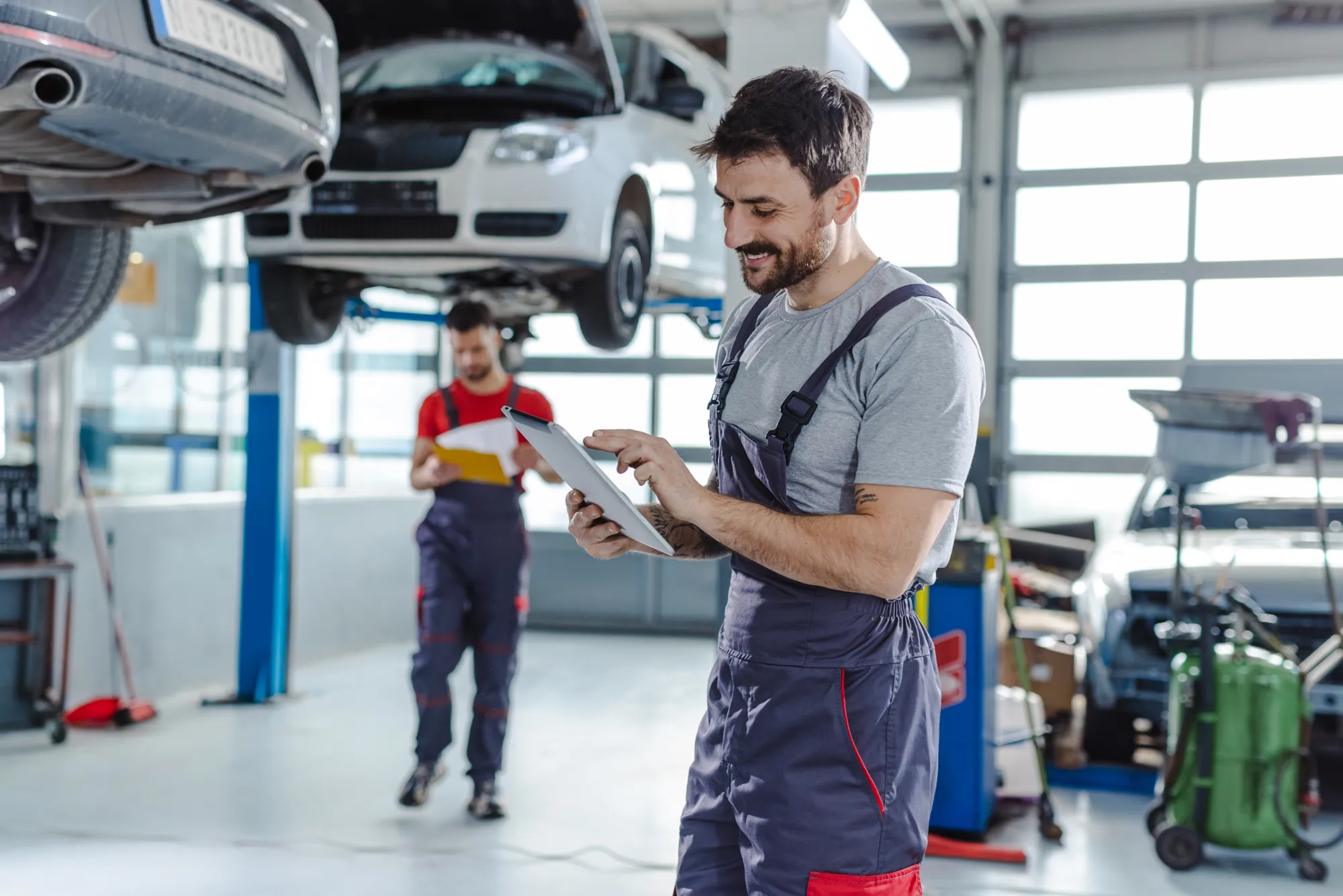 A mechanic in grey overalls looks at a tablet while a coworker with a clipboard stands near a car raised on a lift inside an auto repair shop.