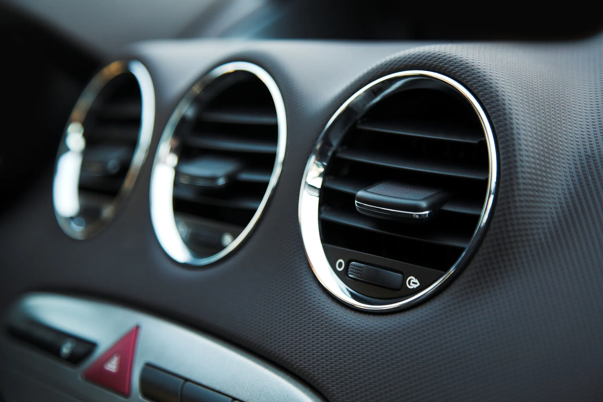 Close-up of three round chrome-trimmed air conditioning vents on a car's dark grey dashboard.